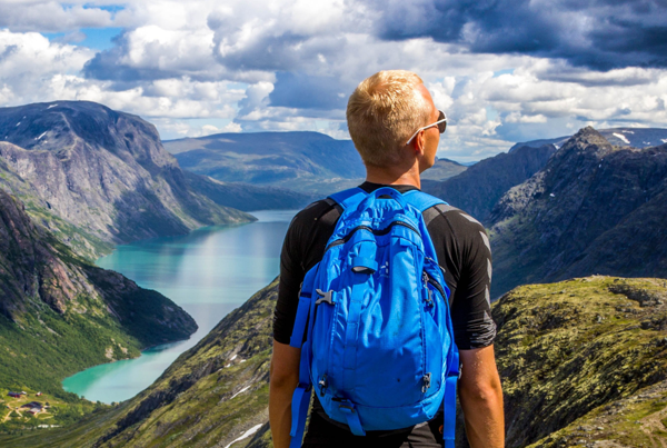 Ein Mann mit Rucksack schaut vom Gipfel ins Tal mit einem See und auf umliegende Gipfel.