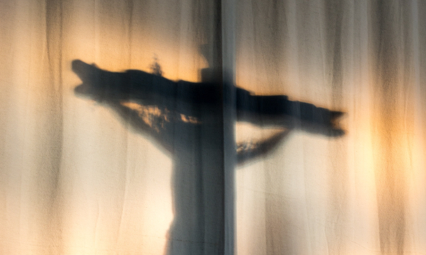 Ein Kruzifix von hinten, angeleuchtet, hinter einem hellen Vorhang.